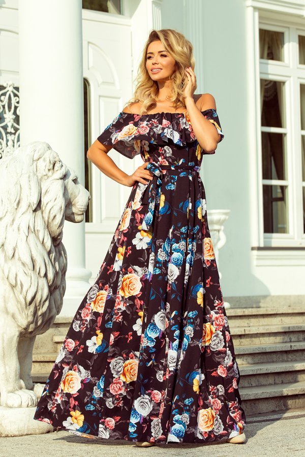 A woman is wearing an elegant, off-the-shoulder floral maxi dress with a vibrant pattern of colorful roses. The dress features a flowing silhouette and a matching belt, perfect for a stylish summer look.