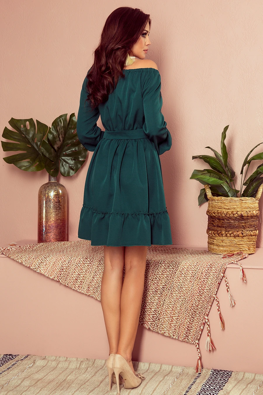 A woman is wearing an elegant, off-the-shoulder teal dress with a ruffled hem and a matching belt. The dress is styled with neutral high heels, creating a sophisticated and chic look.
