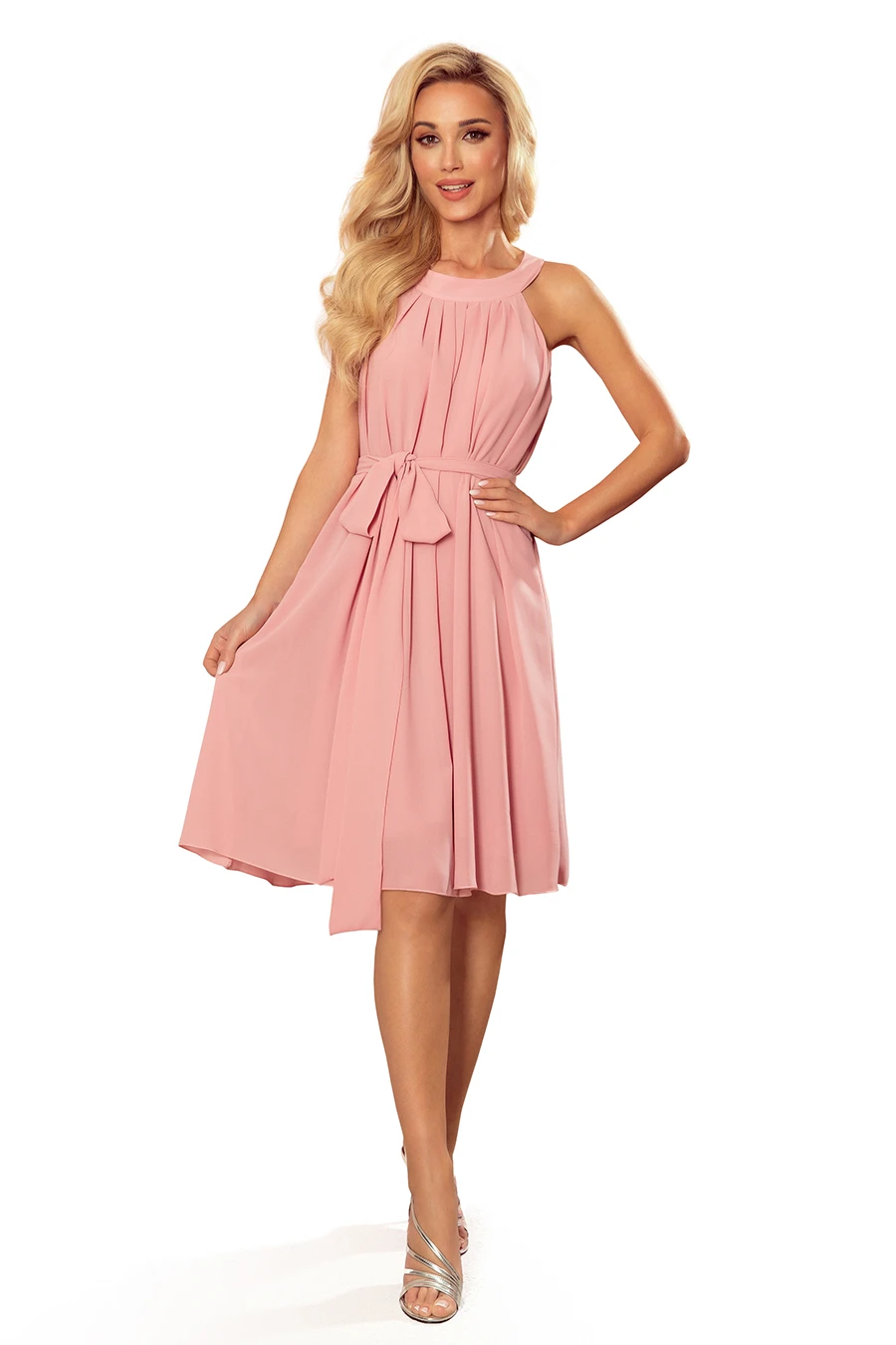 A woman is wearing a sleeveless, knee-length pink dress with a pleated design and a bow at the waist. The dress is paired with silver strappy heels, creating an elegant and stylish look.