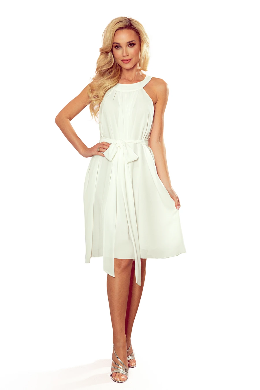 A woman is wearing an elegant, sleeveless white dress with a halter neckline and a waist tie. The dress is paired with strappy silver heels, creating a sophisticated and stylish look.