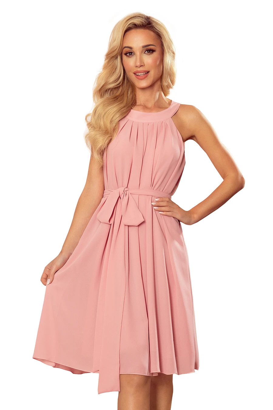 A woman is wearing a sleeveless, knee-length pink dress with a pleated design and a bow at the waist. The dress has a soft, flowing fabric that creates an elegant and feminine look.