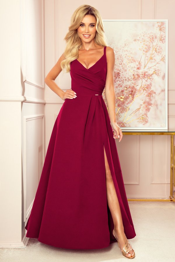 A woman is wearing an elegant, floor-length burgundy gown with a fitted bodice and a stylish slit. The dress features delicate shoulder straps and a flattering, pleated waist design.