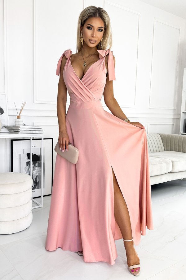 A woman is wearing an elegant pink evening gown with a deep V-neckline and a high slit, complemented by bow details on the shoulders. She holds a beige clutch, adding a touch of sophistication to the ensemble.