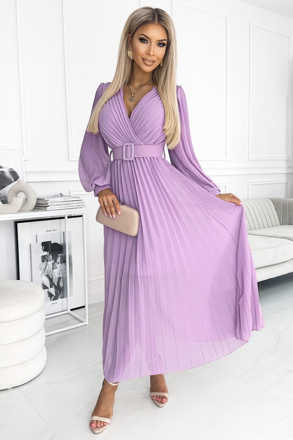 A woman is wearing a flowing, pleated lavender dress with a matching belt, exuding elegance and style. She holds a beige clutch, complementing her sophisticated look in a modern, minimalist setting.