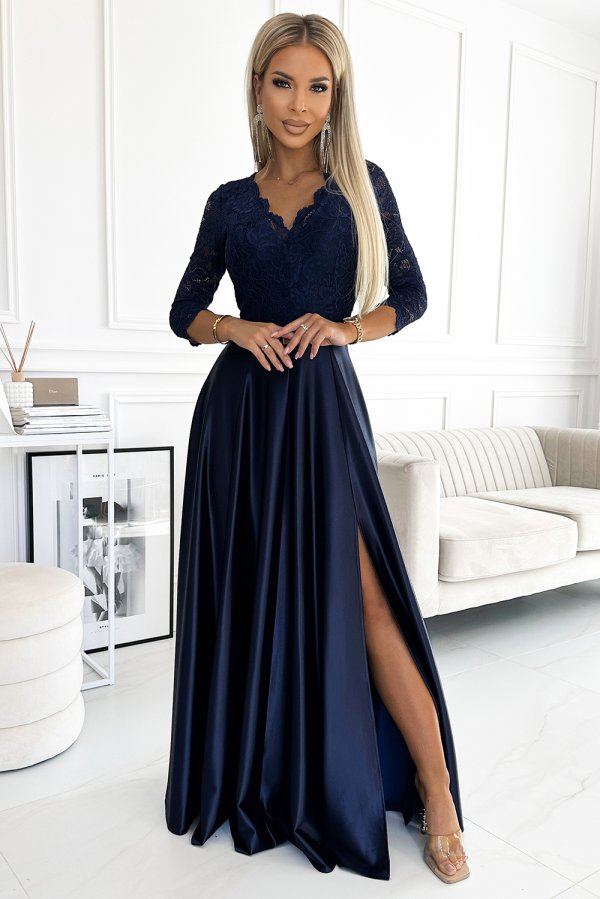 A woman is wearing an elegant navy blue evening gown with a lace bodice and a flowing satin skirt featuring a high slit. The dress is complemented by delicate jewelry, creating a sophisticated and stylish look.