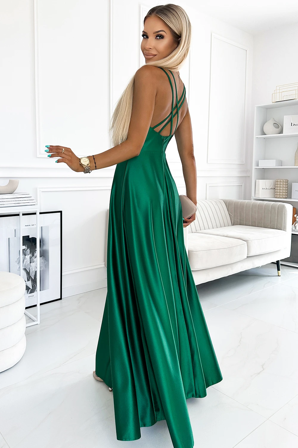 A woman is wearing an elegant, floor-length green dress with thin straps and a flowing skirt. The dress is styled in a modern, minimalist room, highlighting its vibrant color and sophisticated design.
