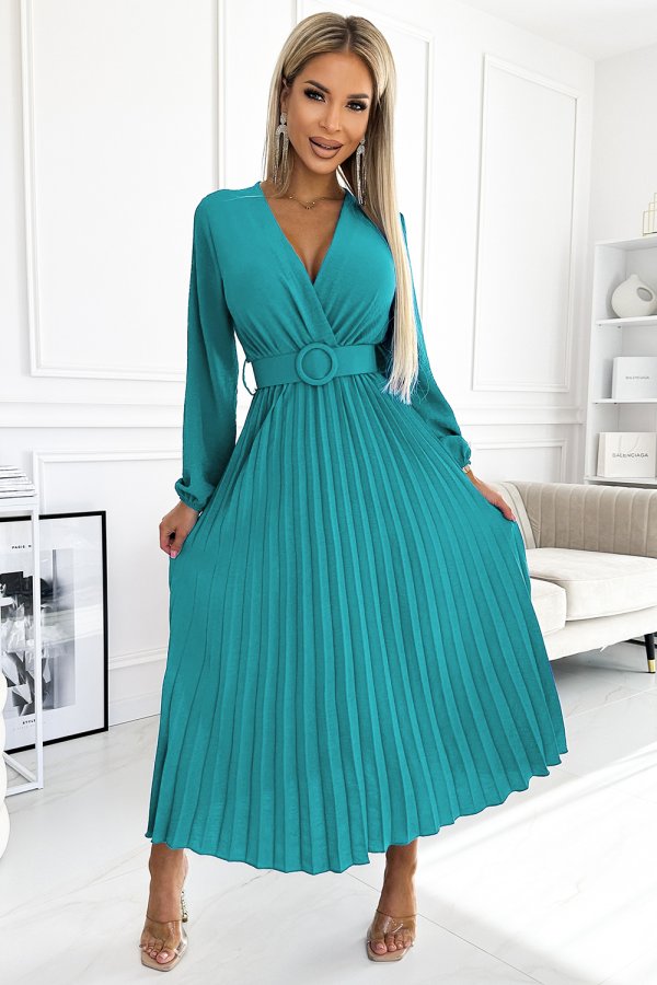A woman is wearing a stylish teal dress with a pleated skirt and a matching belt. The dress features long sleeves and a deep V-neckline, creating an elegant and sophisticated look.