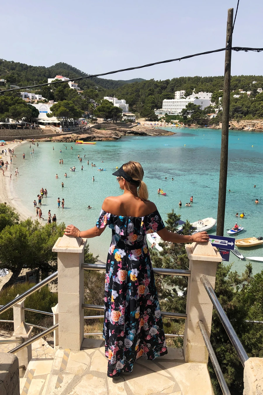A woman is wearing a vibrant floral off-the-shoulder maxi dress, perfect for a summer day by the beach. The dress features a colorful pattern that stands out against the scenic coastal backdrop.