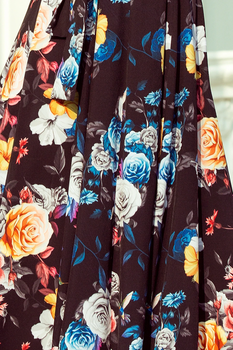 A flowing dress features a vibrant floral pattern with roses in shades of blue, yellow, and white against a dark background. The design exudes elegance and sophistication, perfect for a stylish occasion.