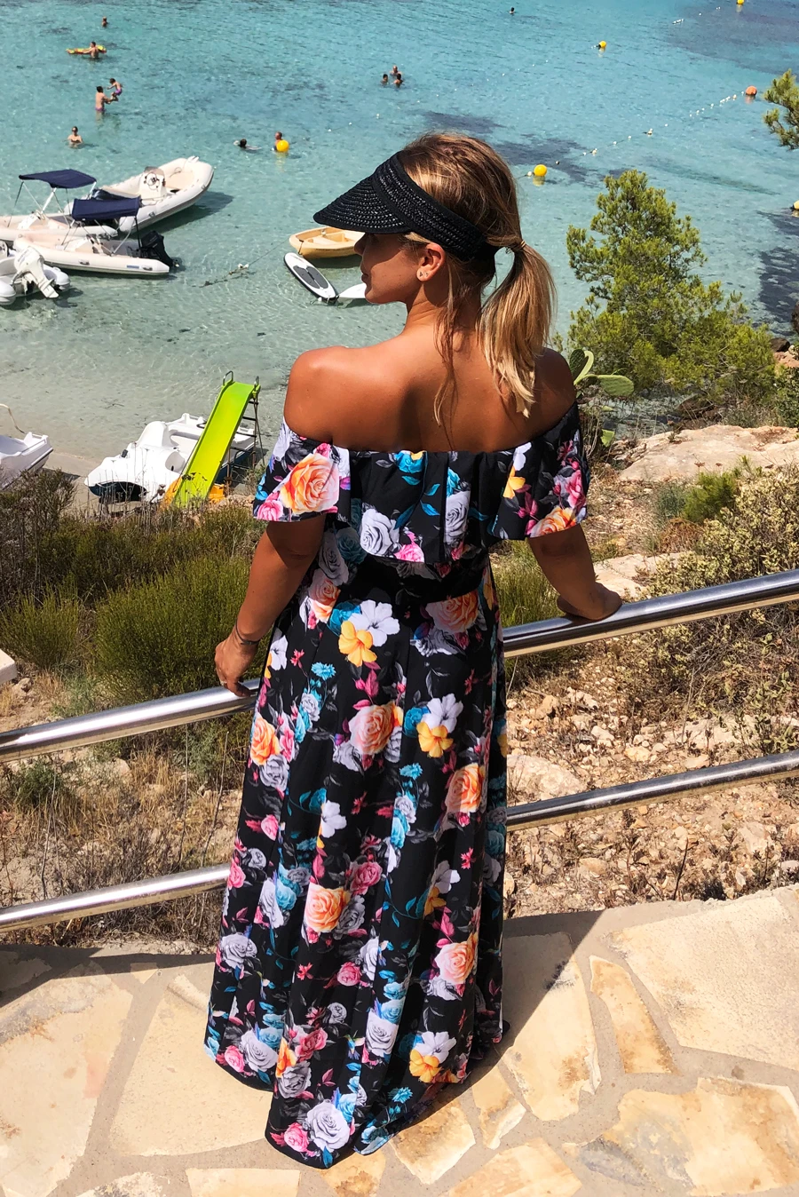A woman is wearing a vibrant, off-the-shoulder floral dress, perfect for a summer day by the sea. The dress features a colorful pattern of roses and other flowers on a dark background, offering a stylish and breezy look.