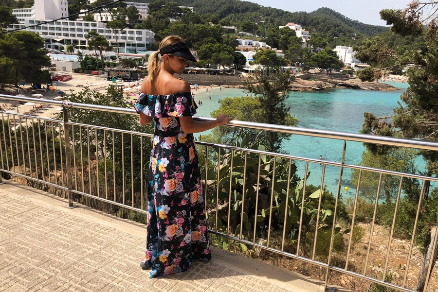 A woman is wearing a vibrant, floral off-the-shoulder maxi dress, perfect for a summer outing. The dress features a colorful pattern that stands out against the scenic coastal backdrop.