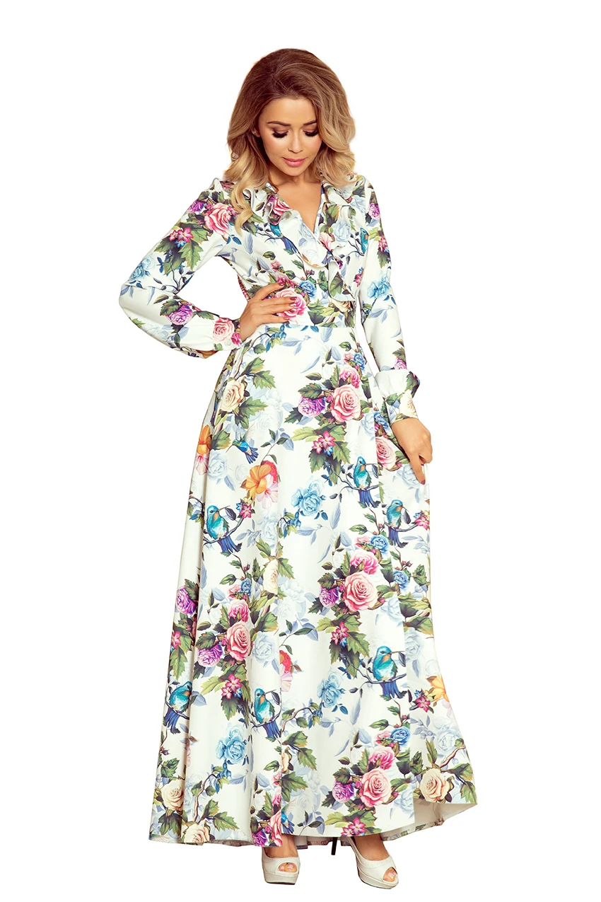 A long-sleeved, full-length dress features a vibrant floral pattern with a mix of colorful flowers and birds on a white background. The dress has a fitted waist and a flowing skirt, creating an elegant and feminine silhouette.
