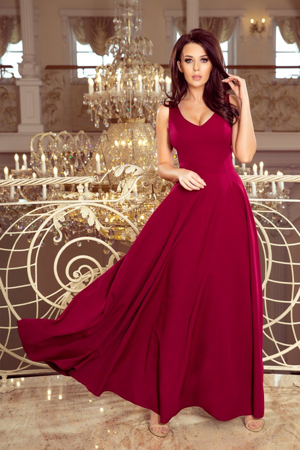 A woman is wearing an elegant, flowing burgundy evening gown with a fitted bodice and a flared skirt. The dress is showcased in a luxurious setting with ornate chandeliers in the background.