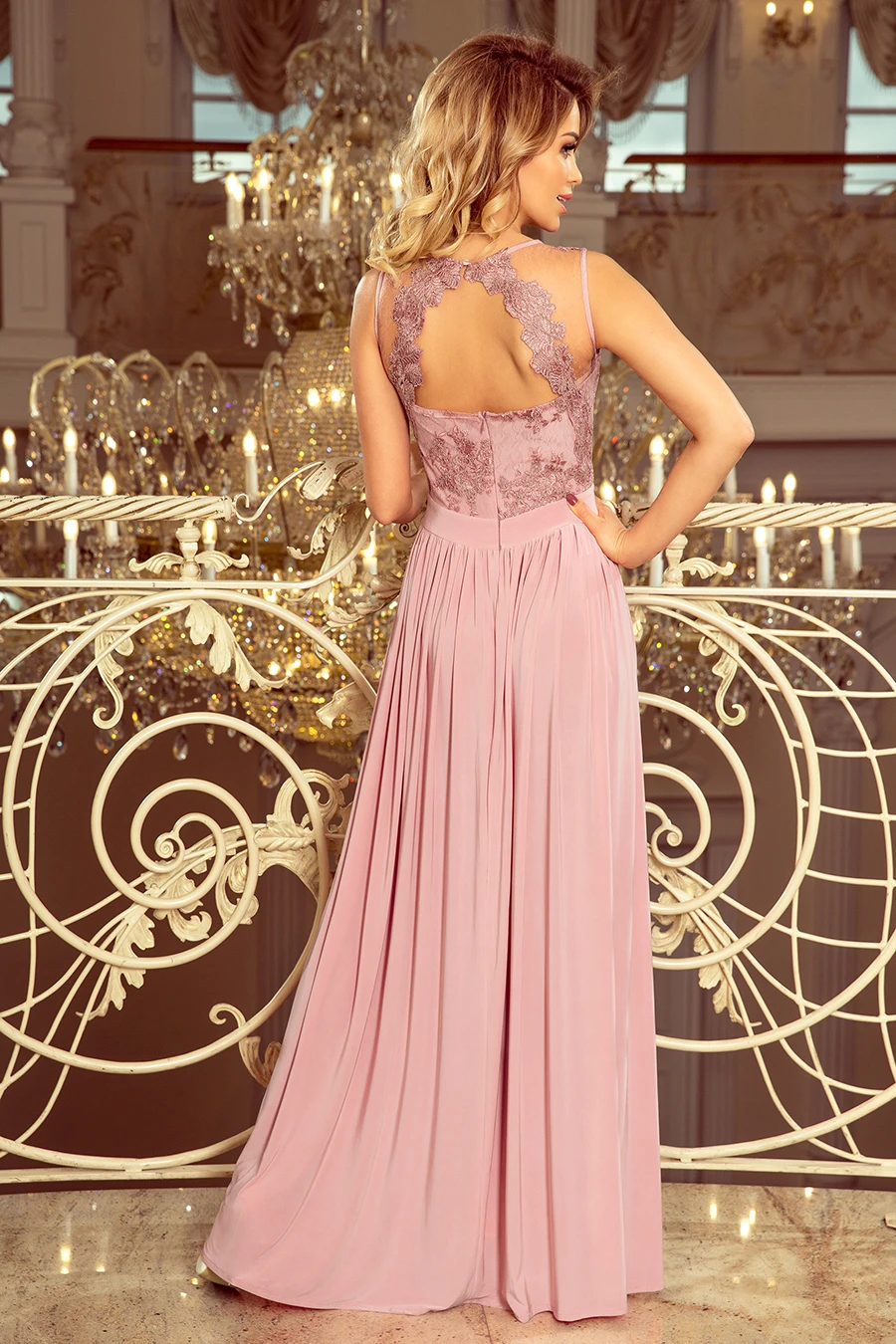 A woman is wearing an elegant, floor-length pink gown with intricate lace detailing on the back. The dress is showcased in a luxurious setting with ornate decor and a grand chandelier in the background.