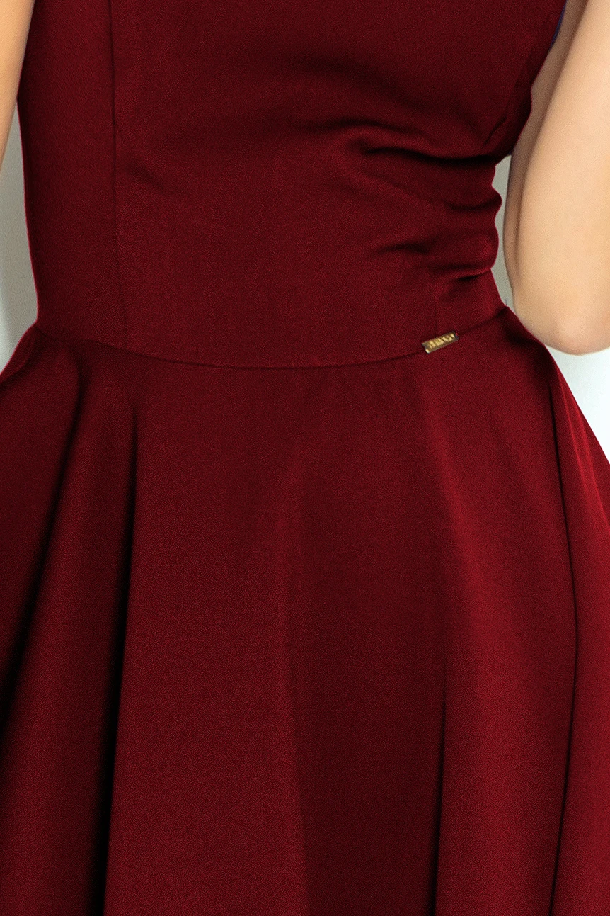 The image shows a close-up of a burgundy dress with a fitted bodice and a flared skirt. The fabric appears smooth and elegant, with a small metallic tag detail at the waist.