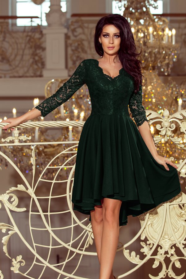 A woman is wearing an elegant dark green dress with lace sleeves and a flowing skirt. The dress is showcased in a luxurious setting with ornate decor and chandeliers in the background.