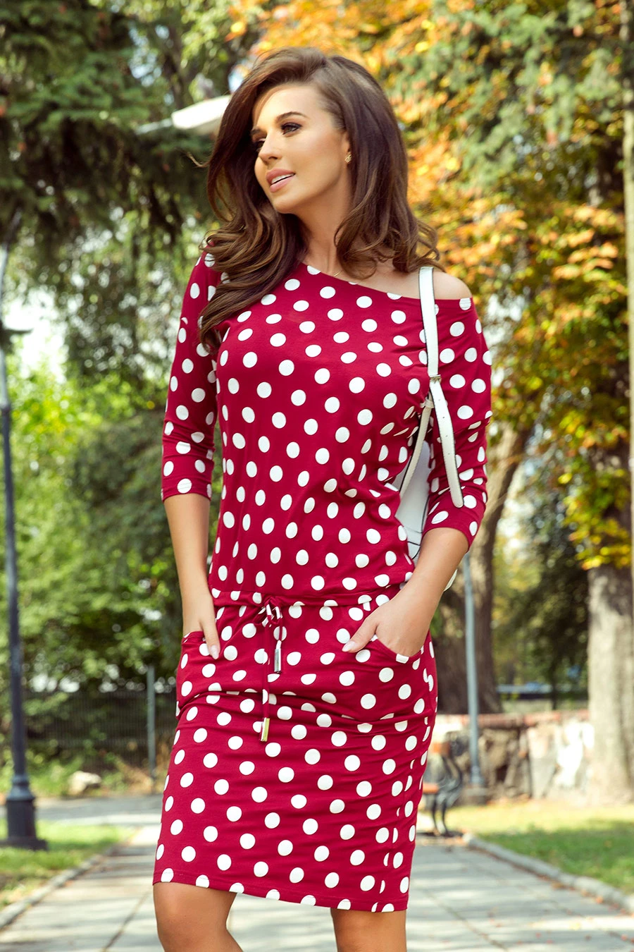 A woman is wearing a stylish red dress with white polka dots, featuring three-quarter sleeves and a drawstring waist. The dress is complemented by a white shoulder bag, creating a chic and casual look.