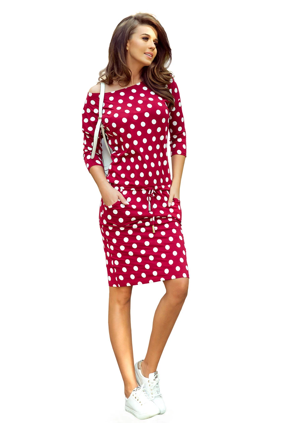 A woman is wearing a stylish red dress with white polka dots, featuring three-quarter sleeves and a drawstring waist. She pairs the dress with white sneakers and carries a white shoulder bag.