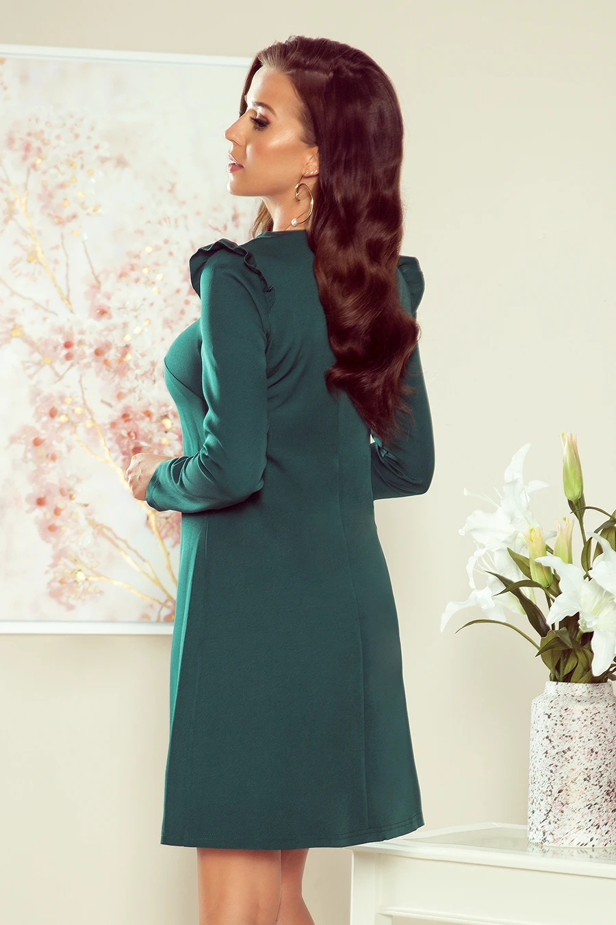 The image features a woman wearing an elegant, long-sleeved green dress with subtle ruffle details on the shoulders. The dress is styled in a sophisticated setting, complemented by a floral painting and a vase of white lilies.