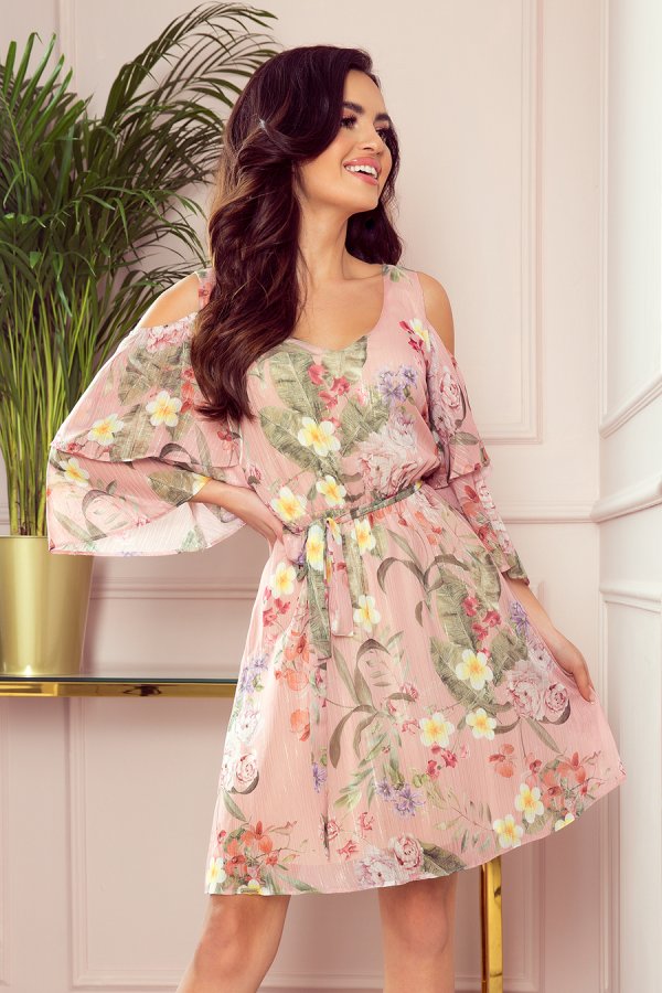 A woman is wearing a pink floral dress with a belted waist and cold shoulder sleeves. The dress features a vibrant pattern of flowers and leaves, creating a fresh and elegant look.