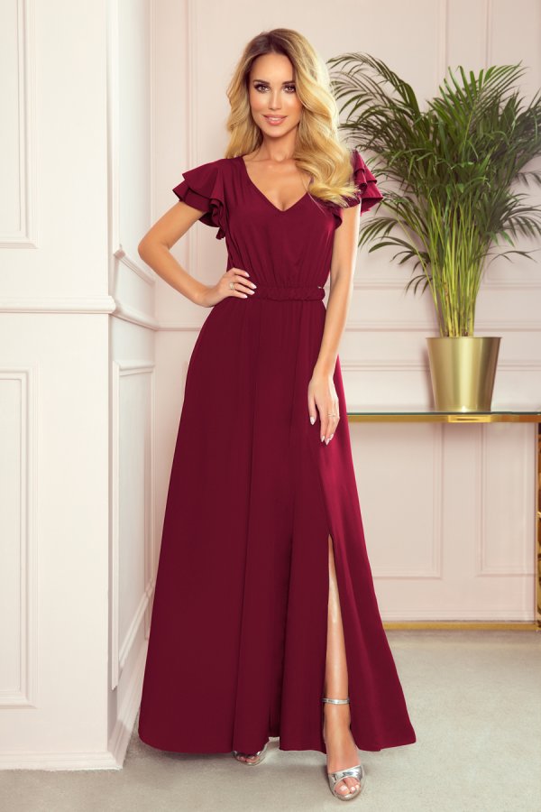 A woman is wearing an elegant, floor-length burgundy dress with ruffled sleeves and a stylish slit. The dress is complemented by silver high-heeled sandals, adding a touch of sophistication.