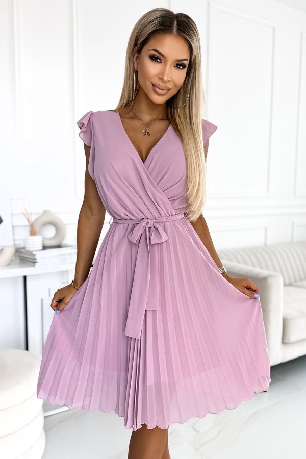 A woman is wearing a stylish, pleated pink dress with a wrap design and a tie at the waist. The dress features delicate cap sleeves and a flowing skirt, perfect for a chic and elegant look.