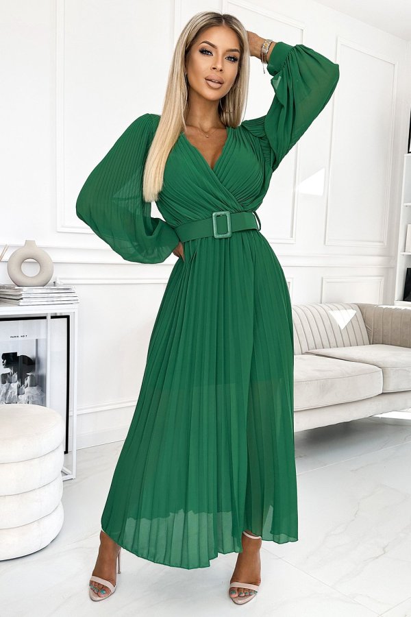 A woman is wearing an elegant green pleated dress with long sleeves and a matching belt. The dress features a deep V-neckline and flows gracefully to the floor.