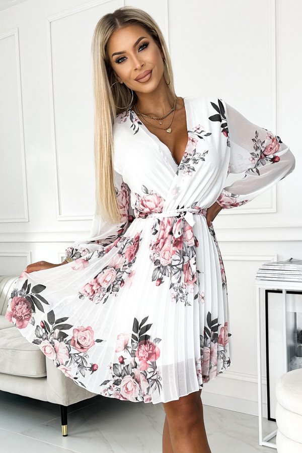 A woman is wearing a white floral dress with pink and black rose patterns, featuring a pleated skirt and a wrap-style top. The dress is elegant and perfect for a spring or summer occasion.