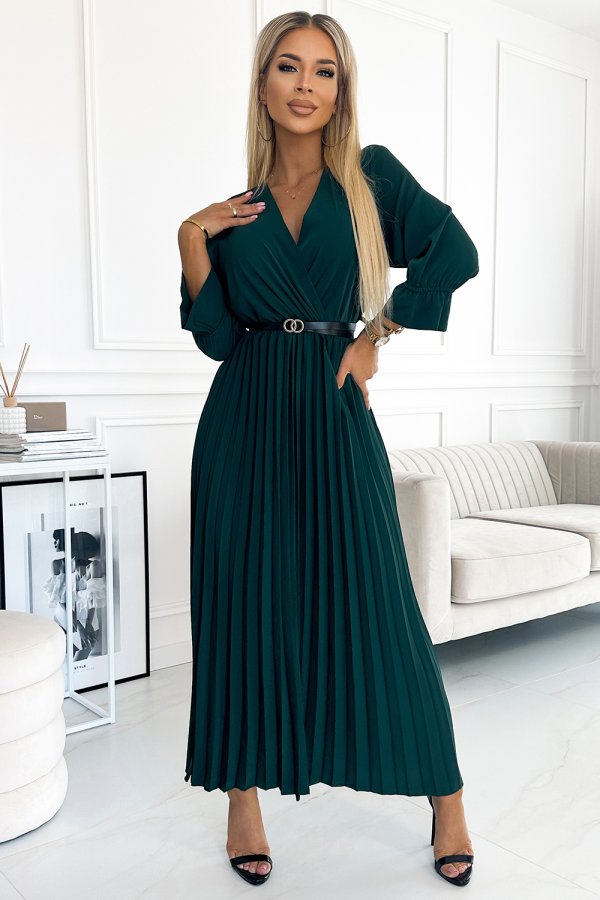 A woman is wearing an elegant, dark green pleated dress with a V-neckline and a black belt accentuating the waist. The dress is paired with black high-heeled sandals, creating a sophisticated and stylish look.