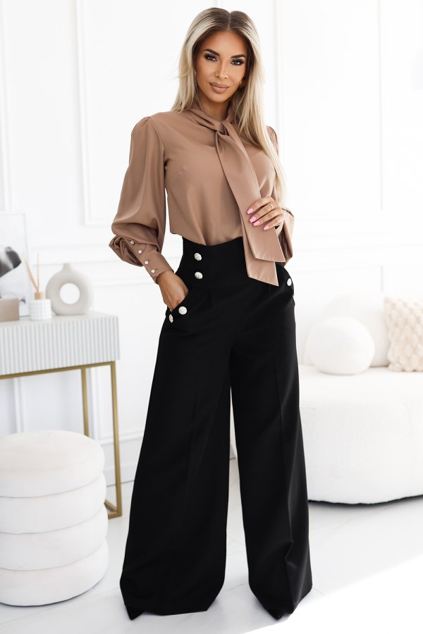 A stylish woman is wearing a chic outfit featuring a beige blouse with a bow tie detail and elegant black high-waisted trousers with decorative buttons. The ensemble exudes sophistication, perfect for a professional or upscale setting.