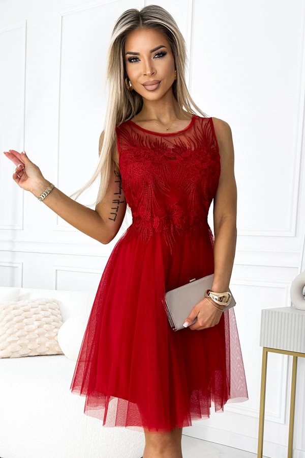 A woman is wearing an elegant red dress with intricate lace detailing and a sheer neckline. She holds a sleek, neutral-colored clutch, complementing the sophisticated look.